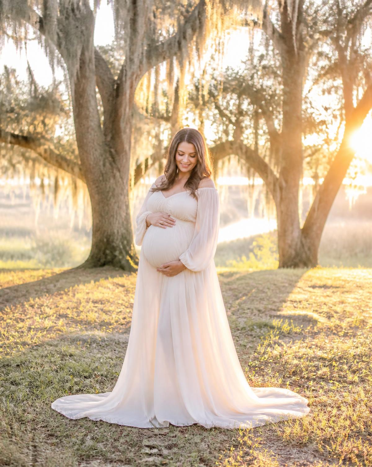 Expectant mother in a flowing cream gown among moss-draped oaks at sunset